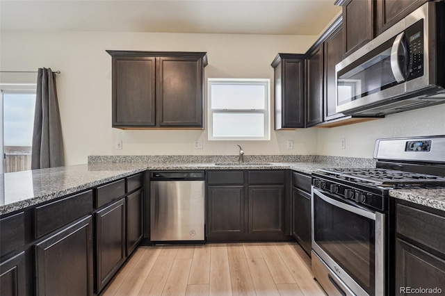 kitchen with appliances with stainless steel finishes, sink, light stone countertops, dark brown cabinets, and light wood-type flooring