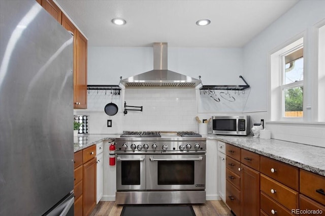 kitchen featuring backsplash, appliances with stainless steel finishes, brown cabinets, wood finished floors, and wall chimney exhaust hood