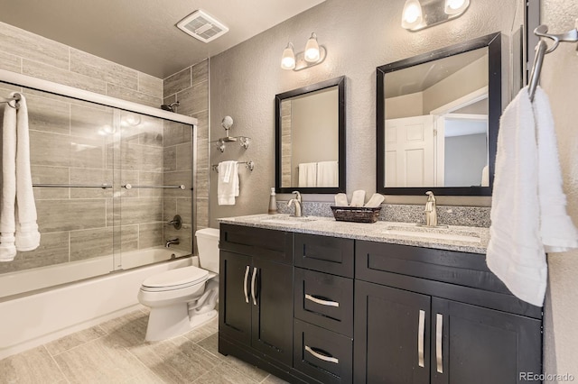 bathroom with a textured wall, toilet, double vanity, and bath / shower combo with glass door