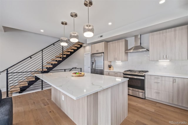 kitchen featuring appliances with stainless steel finishes, hanging light fixtures, a center island, wall chimney exhaust hood, and light wood-type flooring