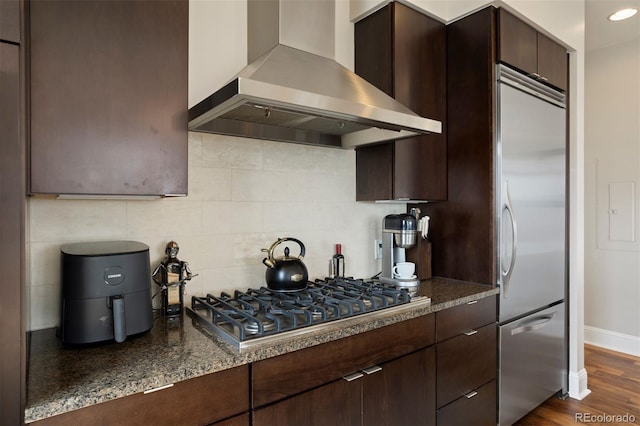 kitchen featuring appliances with stainless steel finishes, backsplash, wall chimney exhaust hood, dark stone countertops, and dark hardwood / wood-style floors