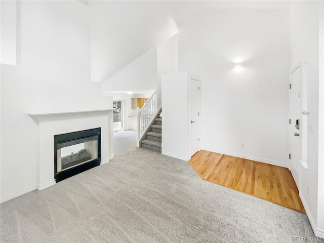 unfurnished living room featuring stairway, high vaulted ceiling, carpet flooring, baseboards, and a glass covered fireplace
