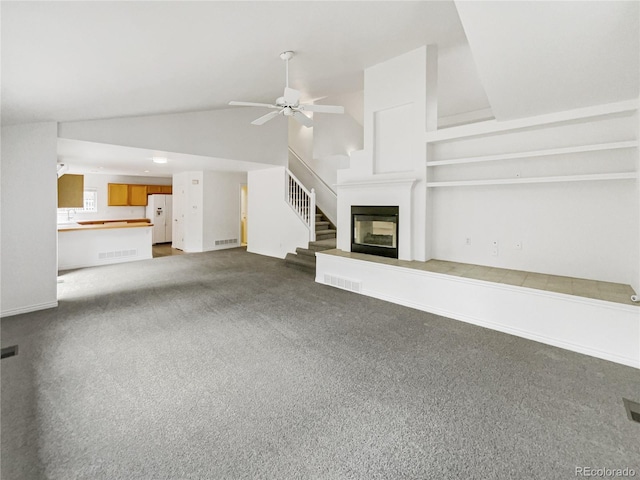 unfurnished living room featuring a ceiling fan, vaulted ceiling, dark colored carpet, a glass covered fireplace, and stairs