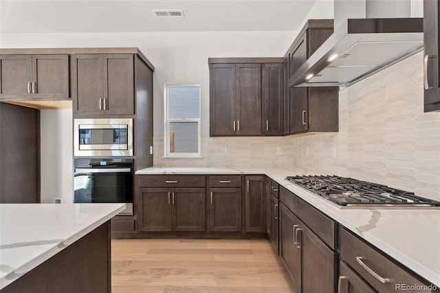 kitchen with wall chimney range hood, light stone counters, stainless steel appliances, and light hardwood / wood-style flooring