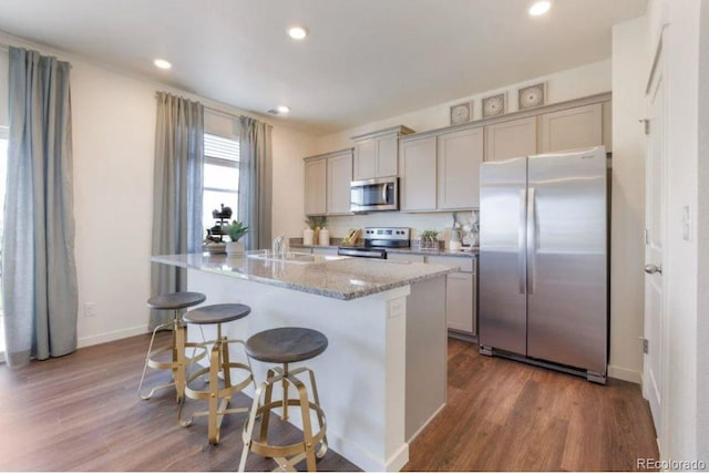 kitchen with hardwood / wood-style floors, light stone counters, stainless steel appliances, gray cabinets, and a kitchen island with sink