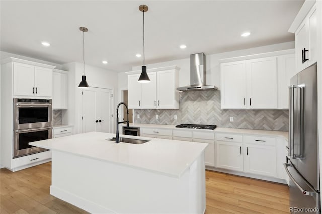 kitchen featuring wall chimney range hood, a center island with sink, hanging light fixtures, and appliances with stainless steel finishes