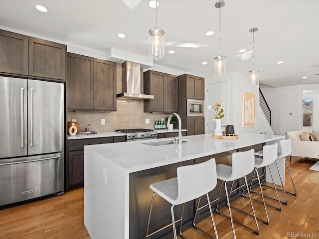 kitchen featuring stainless steel appliances, light wood-style floors, dark brown cabinetry, a sink, and wall chimney exhaust hood