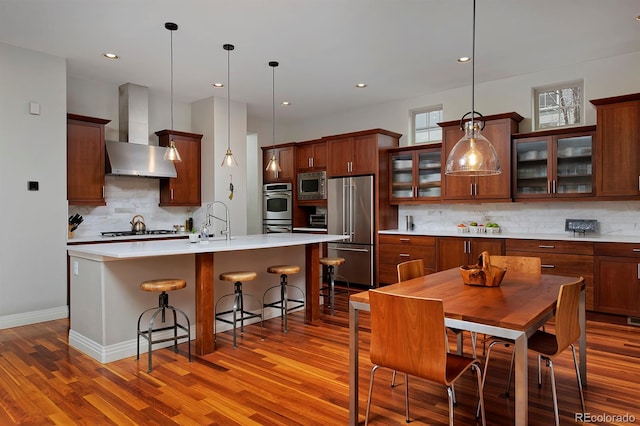 8044 E 24th Ave, Denver, CO 80238 - kitchen featuring appliances with stainless steel finishes, wall chimney range hood, wood finished floors, tasteful backsplash, and a kitchen island with sink