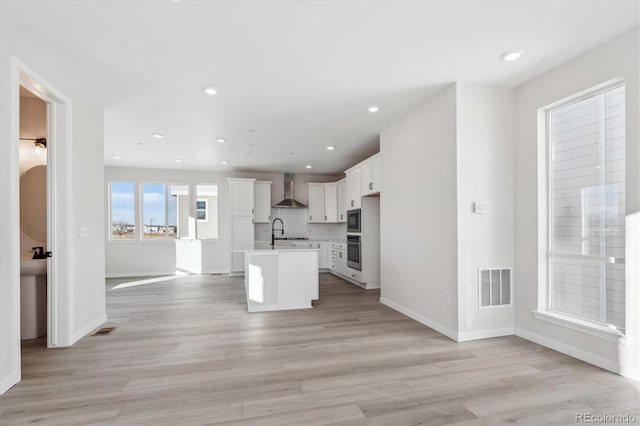 kitchen with white cabinetry, sink, wall chimney range hood, light hardwood / wood-style floors, and a kitchen island with sink