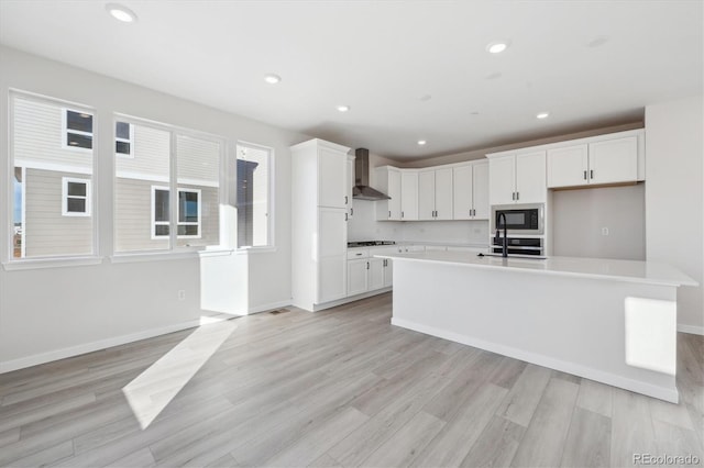 kitchen featuring white cabinets, a center island with sink, black microwave, and wall chimney range hood