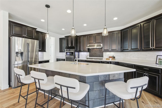 kitchen with an island with sink, under cabinet range hood, a sink, decorative backsplash, and appliances with stainless steel finishes