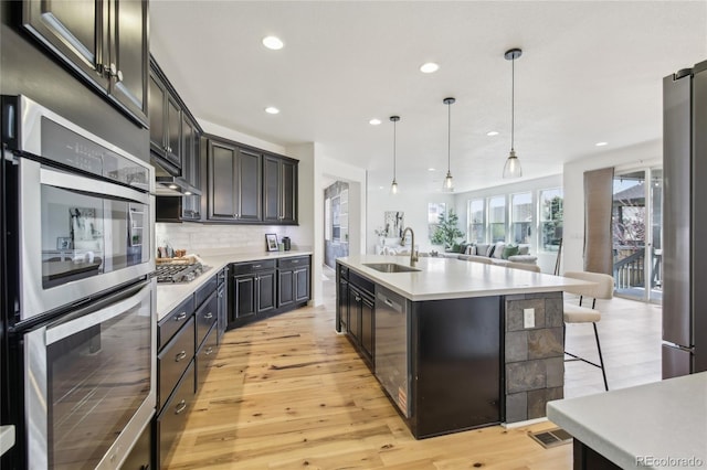 kitchen with a kitchen island with sink, light wood-style flooring, a sink, light countertops, and appliances with stainless steel finishes