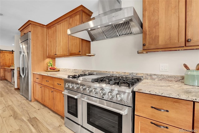 kitchen featuring light stone counters, appliances with stainless steel finishes, wall chimney range hood, light wood finished floors, and brown cabinetry