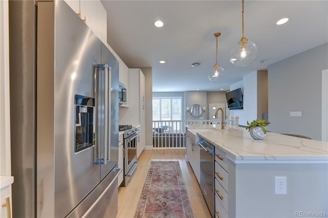 kitchen featuring premium appliances, decorative light fixtures, white cabinets, light stone counters, and a center island with sink