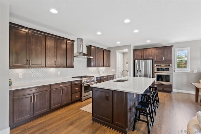 5608 Raintree Dr, Parker, CO 80134 - kitchen featuring wall chimney range hood, appliances with stainless steel finishes, a sink, wood finished floors, and a kitchen breakfast bar