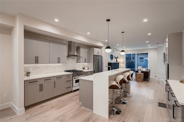 kitchen featuring wall chimney range hood, a kitchen bar, high end appliances, gray cabinetry, and light wood finished floors