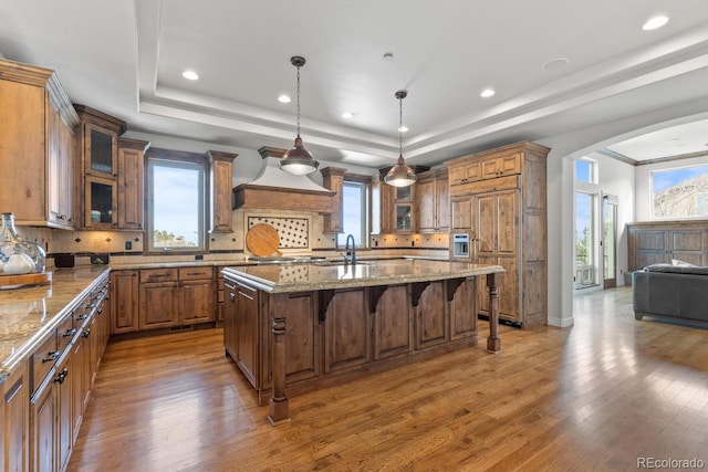 kitchen featuring a breakfast bar, hanging light fixtures, a raised ceiling, light stone countertops, and a kitchen island with sink