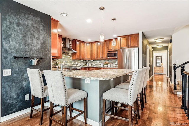 kitchen with wall chimney exhaust hood, stainless steel appliances, backsplash, decorative light fixtures, and hardwood / wood-style flooring