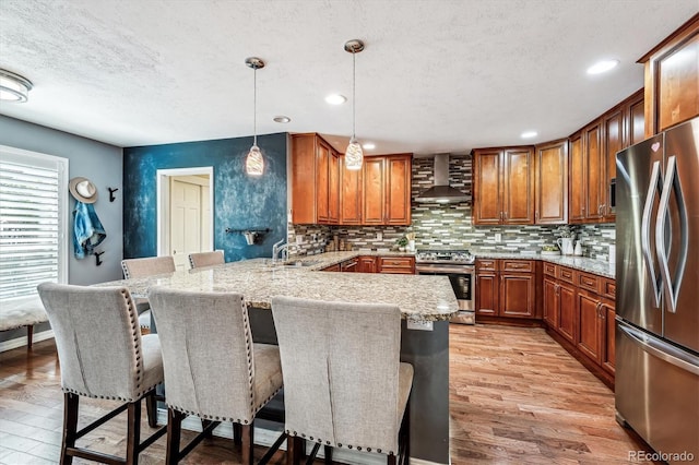 kitchen featuring wall chimney range hood, a kitchen breakfast bar, kitchen peninsula, pendant lighting, and appliances with stainless steel finishes