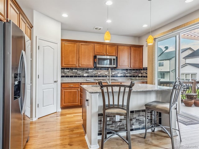 kitchen featuring pendant lighting, light stone counters, appliances with stainless steel finishes, brown cabinetry, and an island with sink