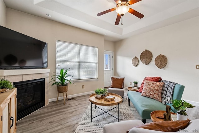 living area featuring ceiling fan, a raised ceiling, light wood-style floors, baseboards, and a tile fireplace