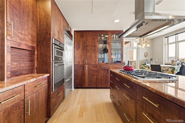 kitchen with stainless steel appliances, light wood-style floors, light stone countertops, island exhaust hood, and glass insert cabinets