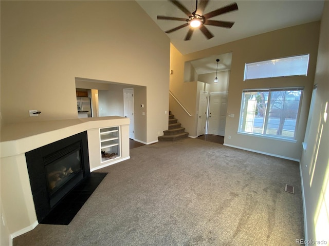 unfurnished living room with ceiling fan, a fireplace with flush hearth, stairs, high vaulted ceiling, and dark colored carpet