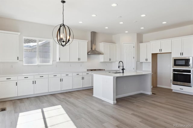 kitchen with stainless steel appliances, an island with sink, white cabinets, decorative light fixtures, and wall chimney exhaust hood