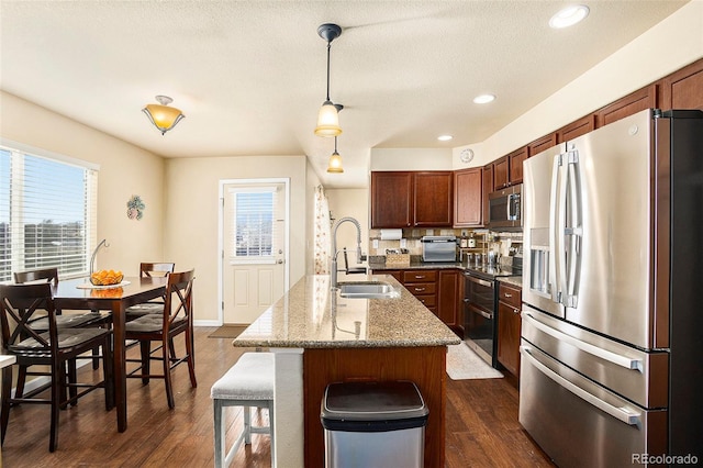 kitchen featuring appliances with stainless steel finishes, dark wood-style flooring, hanging light fixtures, a kitchen island with sink, and a sink