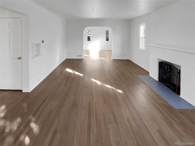 unfurnished living room with arched walkways, a brick fireplace, dark wood finished floors, and baseboards