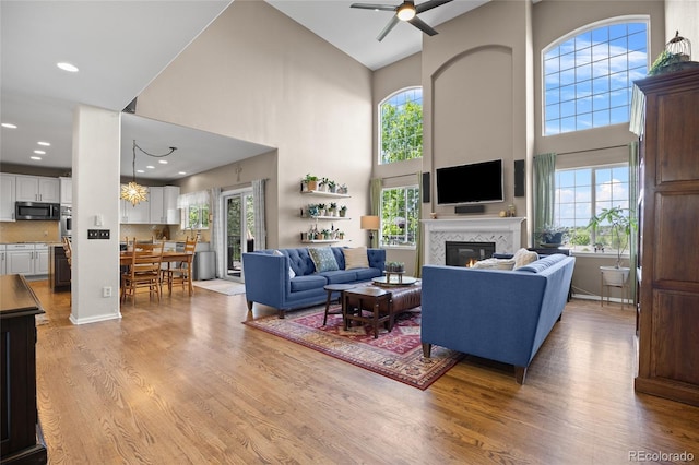 living room featuring a high ceiling, a glass covered fireplace, a ceiling fan, light wood-type flooring, and recessed lighting