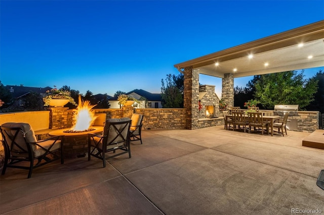 patio terrace at dusk with a grill, an outdoor kitchen, an outdoor stone fireplace, and an outdoor fire pit