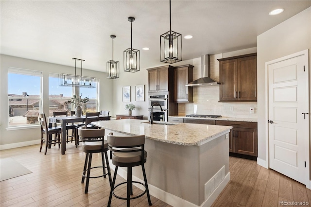 kitchen featuring wall chimney exhaust hood, a sink, stainless steel appliances, an island with sink, and light wood finished floors