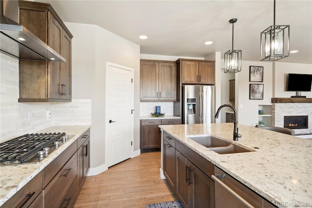 kitchen with appliances with stainless steel finishes, a sink, wall chimney exhaust hood, light wood-style floors, and hanging light fixtures
