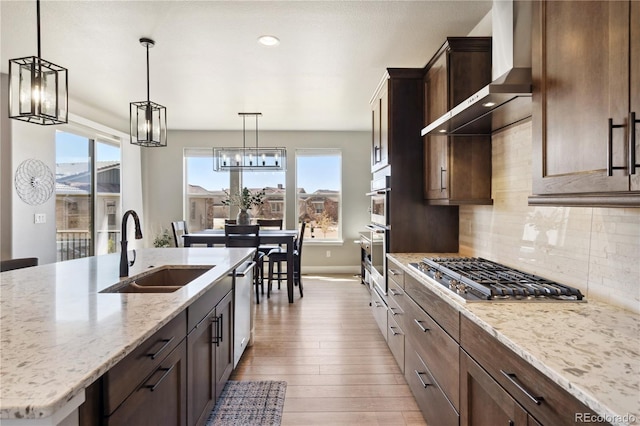 kitchen with a sink, wall chimney range hood, appliances with stainless steel finishes, light wood-style floors, and backsplash