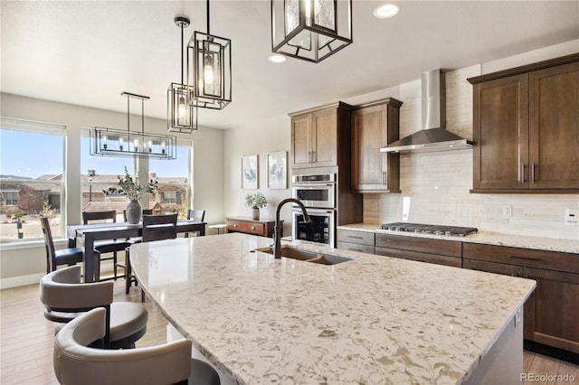 kitchen with wall chimney exhaust hood, stainless steel appliances, light wood-style floors, an island with sink, and a sink