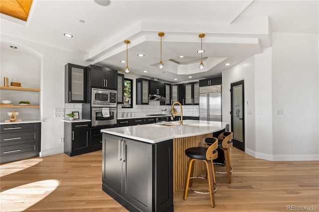 kitchen featuring a kitchen island with sink, hanging light fixtures, built in appliances, decorative backsplash, and light wood-type flooring