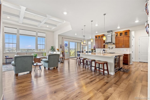 kitchen featuring pendant lighting, wall chimney range hood, light hardwood / wood-style flooring, a kitchen island with sink, and decorative backsplash