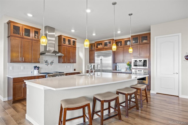 kitchen with a large island, wall chimney range hood, sink, built in appliances, and decorative light fixtures