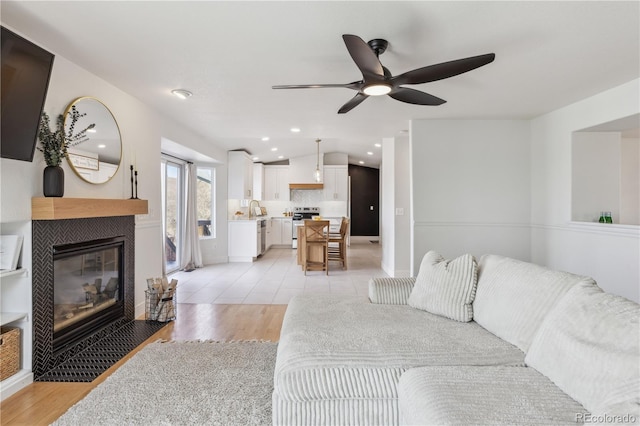 living area featuring a tile fireplace, light wood-style flooring, a ceiling fan, baseboards, and recessed lighting