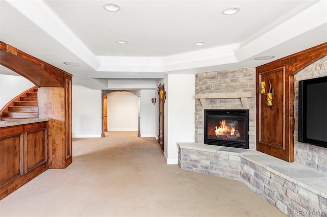unfurnished living room featuring light carpet, a fireplace, arched walkways, baseboards, and a tray ceiling