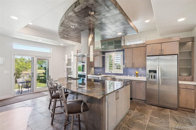 kitchen with dark stone countertops, island exhaust hood, a kitchen breakfast bar, appliances with stainless steel finishes, and a center island with sink