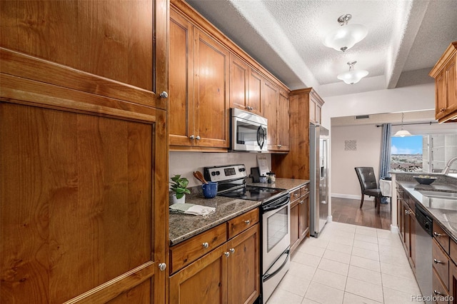 kitchen with appliances with stainless steel finishes, light tile patterned flooring, brown cabinetry, a textured ceiling, and a sink