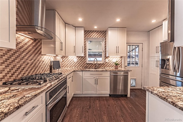 kitchen with light stone counters, a sink, stainless steel appliances, white cabinets, and wall chimney range hood