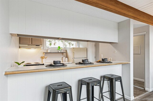 kitchen with a kitchen bar, wood finished floors, freestanding refrigerator, a peninsula, and under cabinet range hood