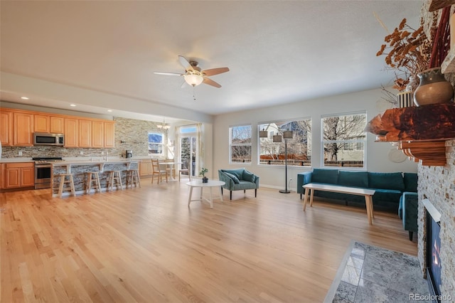 living room with light wood-type flooring, ceiling fan with notable chandelier, plenty of natural light, and a stone fireplace