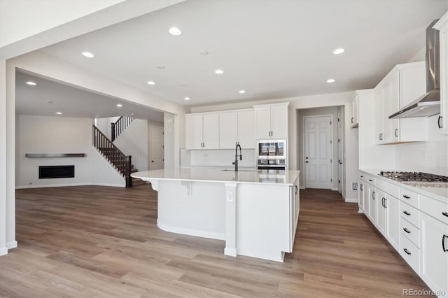 kitchen featuring white cabinetry, stainless steel gas stovetop, a kitchen island with sink, light hardwood / wood-style flooring, and wall chimney range hood