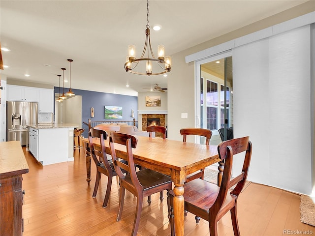 dining space featuring light wood finished floors, a lit fireplace, ceiling fan with notable chandelier, and recessed lighting