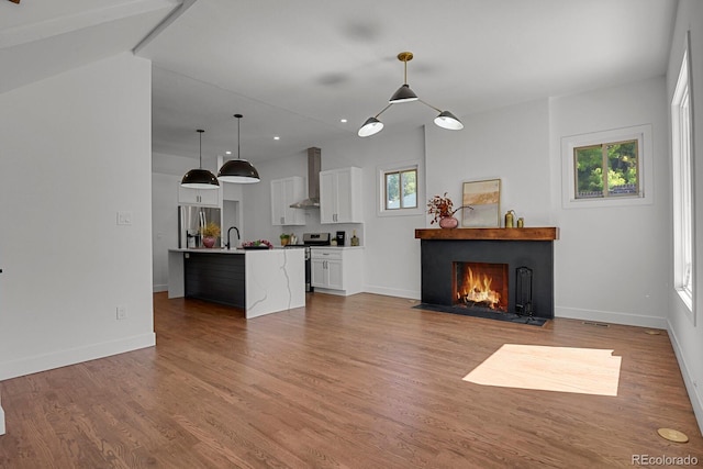 interior space with an island with sink, decorative light fixtures, wall chimney exhaust hood, white cabinetry, and stainless steel appliances