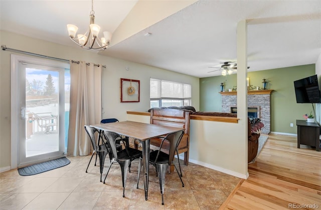 tiled dining room with ceiling fan with notable chandelier, a stone fireplace, and vaulted ceiling